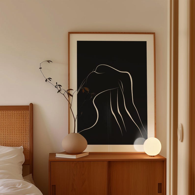 May include: A black and white line art print of a figure in a minimalist style, framed in a light wood frame, hanging on a wall above a wooden dresser with a white lamp on the right side.