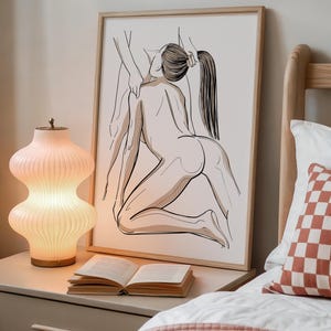 Może przedstawiać: Oprawiony wydruk grafiki liniowej przedstawia nagą postać w pozycji klęczącej, z widocznymi dłońmi innej postaci. Grafika jest w jasnej drewnianej ramie, na stoliku nocnym z lampą i otwartą książką. W tle ł&oacute;żko z poduszką w kratę.