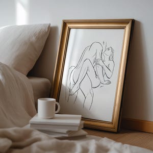 May include: A framed art print featuring a black line drawing of two figures in an embrace. The artwork is in a gold-colored frame and leans against a wall. A white mug sits on a stack of books.