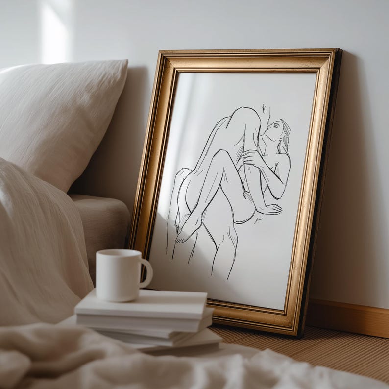 May include: A framed art print featuring a black line drawing of two figures in an embrace. The artwork is in a gold-colored frame and leans against a wall. A white mug sits on a stack of books.