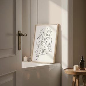 May include: A framed art print featuring a minimalist line drawing of two figures embracing. The artwork is in black against a white background, set within a light wood frame. The print is displayed on a white surface, with candles and a small table nearby.