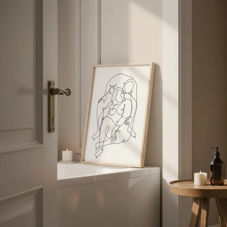 May include: A framed art print featuring a minimalist line drawing of two figures embracing. The artwork is in black against a white background, set within a light wood frame. The print is displayed on a white surface, with candles and a small table nearby.