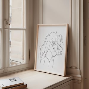 May include: A framed black and white line art print depicting two figures in an embrace. The artwork is displayed on a light-colored wooden surface near a window. The frame is a light wood color.