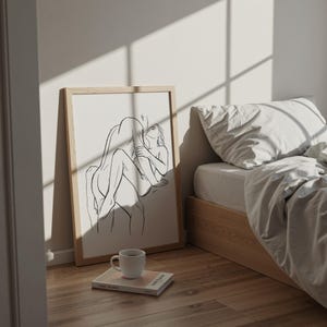 May include: A framed art print featuring a black line drawing of two figures embracing. The artwork is displayed on a wooden floor next to a bed with white linens. A white coffee mug and a book are in the foreground.