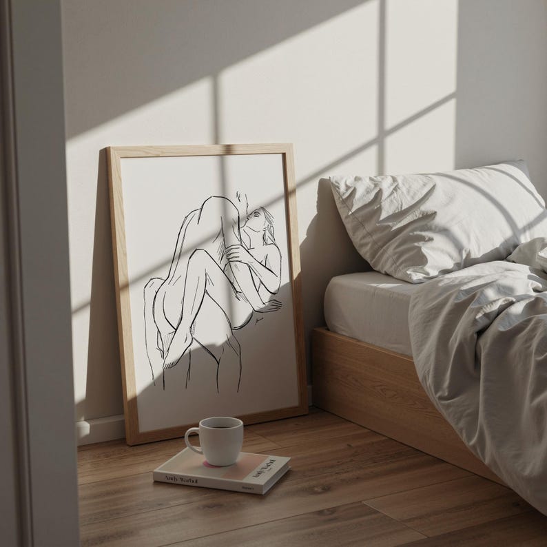 May include: A framed art print featuring a black line drawing of two figures embracing. The artwork is displayed on a wooden floor next to a bed with white linens. A white coffee mug and a book are in the foreground.