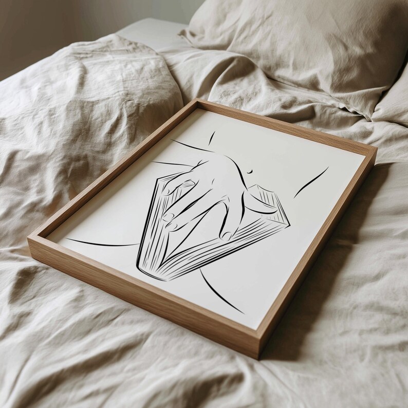 May include: A framed art print featuring a minimalist line drawing of a hand resting on an open book. The artwork is in black and white, set against a white background, and framed in natural wood.