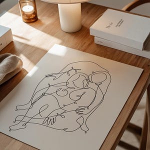 May include: A black line art print of two figures embracing, set on a cream-colored background. The artwork is displayed on a wooden table, alongside a lamp, books, and a candle in a glass holder. The print's minimalist style adds a touch of modern art.