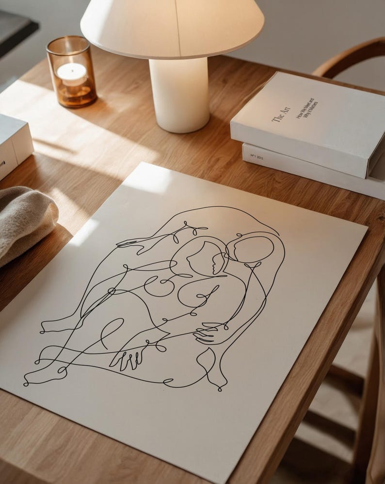 May include: A black line art print of two figures embracing, set on a cream-colored background. The artwork is displayed on a wooden table, alongside a lamp, books, and a candle in a glass holder. The print's minimalist style adds a touch of modern art.