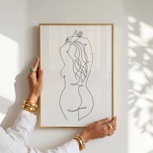 Puede incluir: Una impresi&oacute;n art&iacute;stica enmarcada con un dibujo lineal minimalista de la espalda de una persona. La obra de arte es una l&iacute;nea negra continua sobre un fondo blanco, en un marco dorado. Los brazos de la persona est&aacute;n levantados y el pelo fluye.