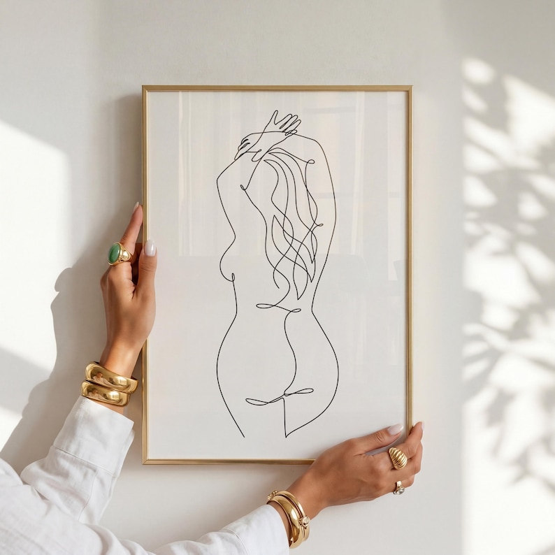 Puede incluir: Una impresi&oacute;n art&iacute;stica enmarcada con un dibujo lineal minimalista de la espalda de una persona. La obra de arte es una l&iacute;nea negra continua sobre un fondo blanco, en un marco dorado. Los brazos de la persona est&aacute;n levantados y el pelo fluye.