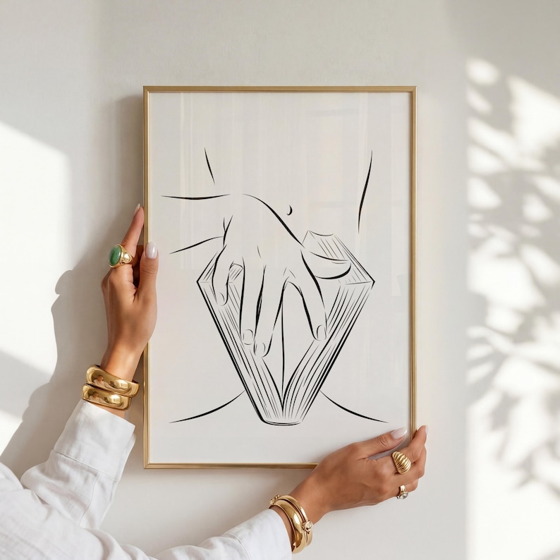 May include: A framed line art print featuring a minimalist illustration of a hand holding open a book. The artwork is in black lines on a white background, set within a gold frame. The print is held by a person wearing gold jewelry.