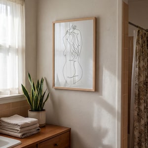 Puede incluir: Una impresi&oacute;n art&iacute;stica enmarcada con un dibujo lineal minimalista de la silueta de una persona. La obra de arte es en negro sobre un fondo blanco, encerrada en un marco de madera clara. La imagen se muestra en un ba&ntilde;o, con una planta y toallas visibles.