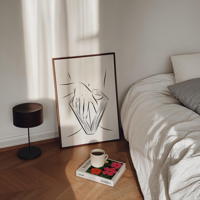May include: A framed black and white line art print of a hand holding a book, leaning against a wall. A small black lamp and a coffee cup on a book with colorful flowers are on the floor. The bed has white linens.