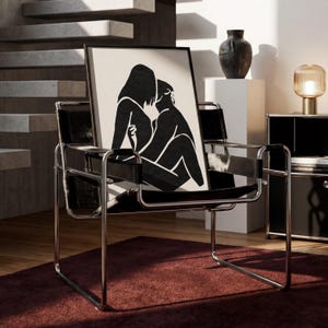 May include: A black and white framed art print featuring a minimalist illustration of two figures embracing. The artwork is displayed on a black leather chair with a chrome frame, set against a neutral-toned interior with a red rug.