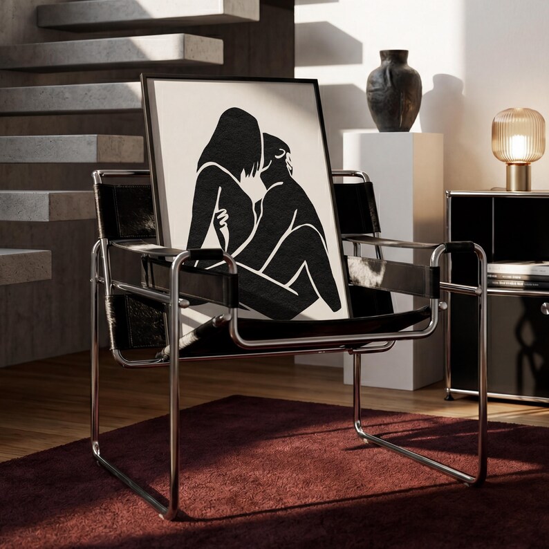May include: A black and white framed art print featuring a minimalist illustration of two figures embracing. The artwork is displayed on a black leather chair with a chrome frame, set against a neutral-toned interior with a red rug.
