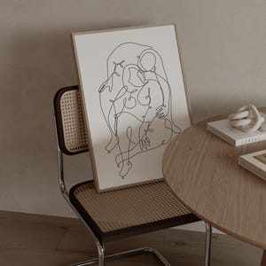 May include: A framed art print featuring a continuous line drawing of two figures embracing. The artwork is in a light-colored wooden frame, leaning against a chair with a woven back. A small, round wooden table holds books and a decorative knot.