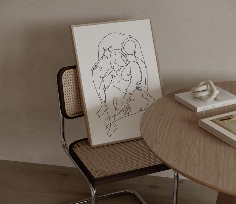 May include: A framed art print featuring a continuous line drawing of two figures embracing. The artwork is in a light-colored wooden frame, leaning against a chair with a woven back. A small, round wooden table holds books and a decorative knot.
