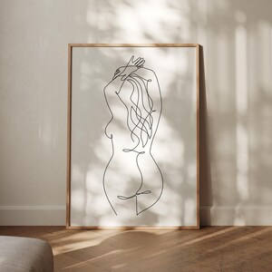 Puede incluir: Una impresi&oacute;n art&iacute;stica enmarcada con un dibujo lineal minimalista de la espalda de una persona. El arte lineal negro representa la forma de la figura, con los brazos levantados hacia la cabeza y el pelo suelto. La obra de arte est&aacute; sobre un fondo neutro.