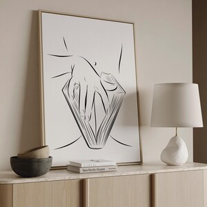 May include: A framed black and white line art print depicts a hand holding a book. The artwork is displayed on a light-colored wall above a wooden cabinet. A white lamp and two decorative bowls are also visible.