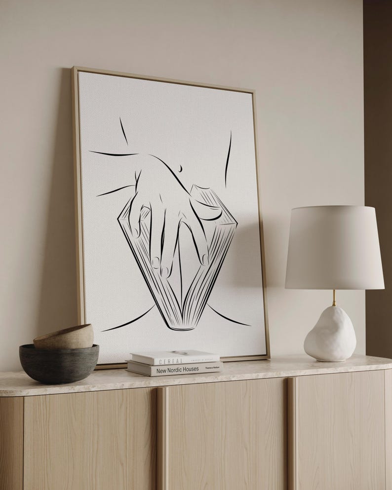 May include: A framed black and white line art print depicts a hand holding a book. The artwork is displayed on a light-colored wall above a wooden cabinet. A white lamp and two decorative bowls are also visible.