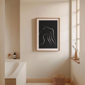 May include: A black and white line art print of a person's body in a relaxed pose, framed in a light wood frame and hanging on a beige wall.