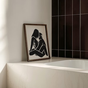 May include: A framed black and white art print depicting two figures in an embrace. The artwork is displayed on a white countertop next to a white bathtub. The frame is a dark brown color.