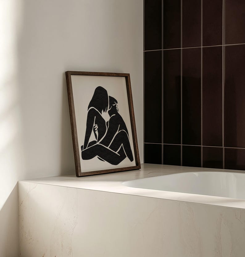 May include: A framed black and white art print depicting two figures in an embrace. The artwork is displayed on a white countertop next to a white bathtub. The frame is a dark brown color.