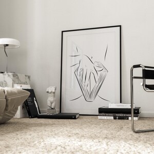 May include: A black framed art print featuring a minimalist line drawing of a hand holding a book. The artwork is displayed on a white wall, alongside books, a marble lamp, and a small sculpture. The room has a neutral color palette.