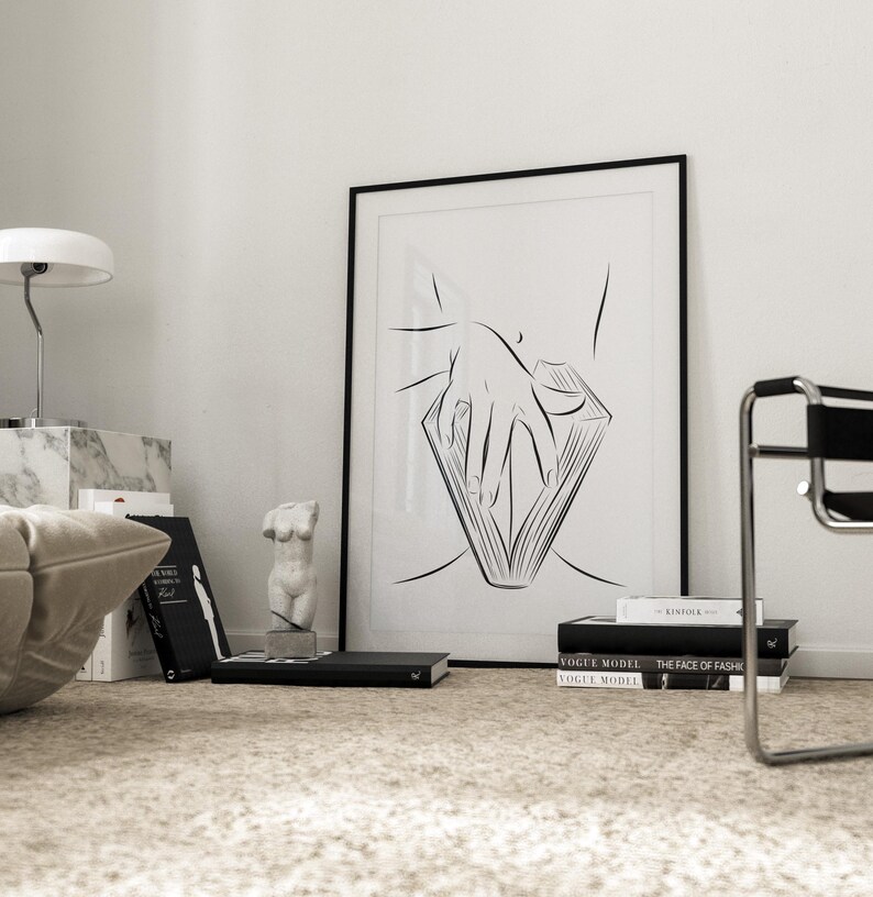 May include: A black framed art print featuring a minimalist line drawing of a hand holding a book. The artwork is displayed on a white wall, alongside books, a marble lamp, and a small sculpture. The room has a neutral color palette.