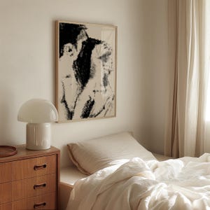 Puede incluir: Una escena de dormitorio con una impresi&oacute;n art&iacute;stica abstracta enmarcada en tonos negros y beige. Una l&aacute;mpara blanca con forma de hongo est&aacute; sobre una c&oacute;moda de madera. La cama est&aacute; hecha con s&aacute;banas blancas y una cortina beige a la derecha.