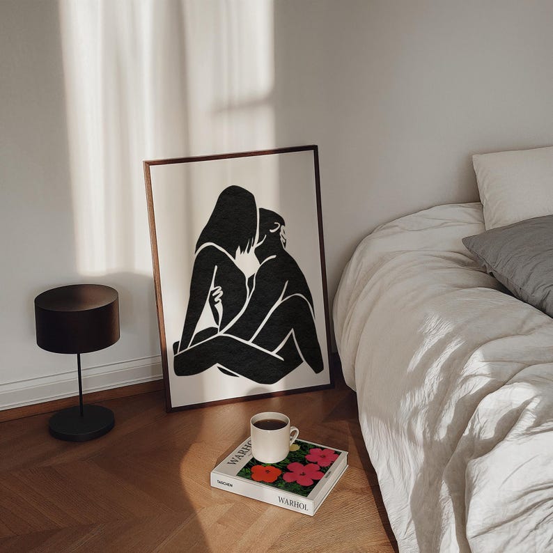 May include: A framed black and white art print depicting two figures embracing. The artwork leans against a wall near a bed with white linens. A coffee cup sits on a book with colorful floral art.