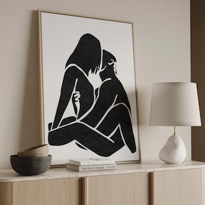 May include: Black and white abstract art print depicting two figures embracing. The artwork is framed in a light wood frame and displayed on a light-colored cabinet. A white lamp and decorative bowls are also visible.