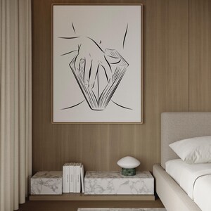May include: A framed black and white line art print depicting a hand holding an open book. The artwork hangs above a marble-topped nightstand with books and a mushroom-shaped lamp. The room has a neutral color palette with wood paneling and a beige bed.