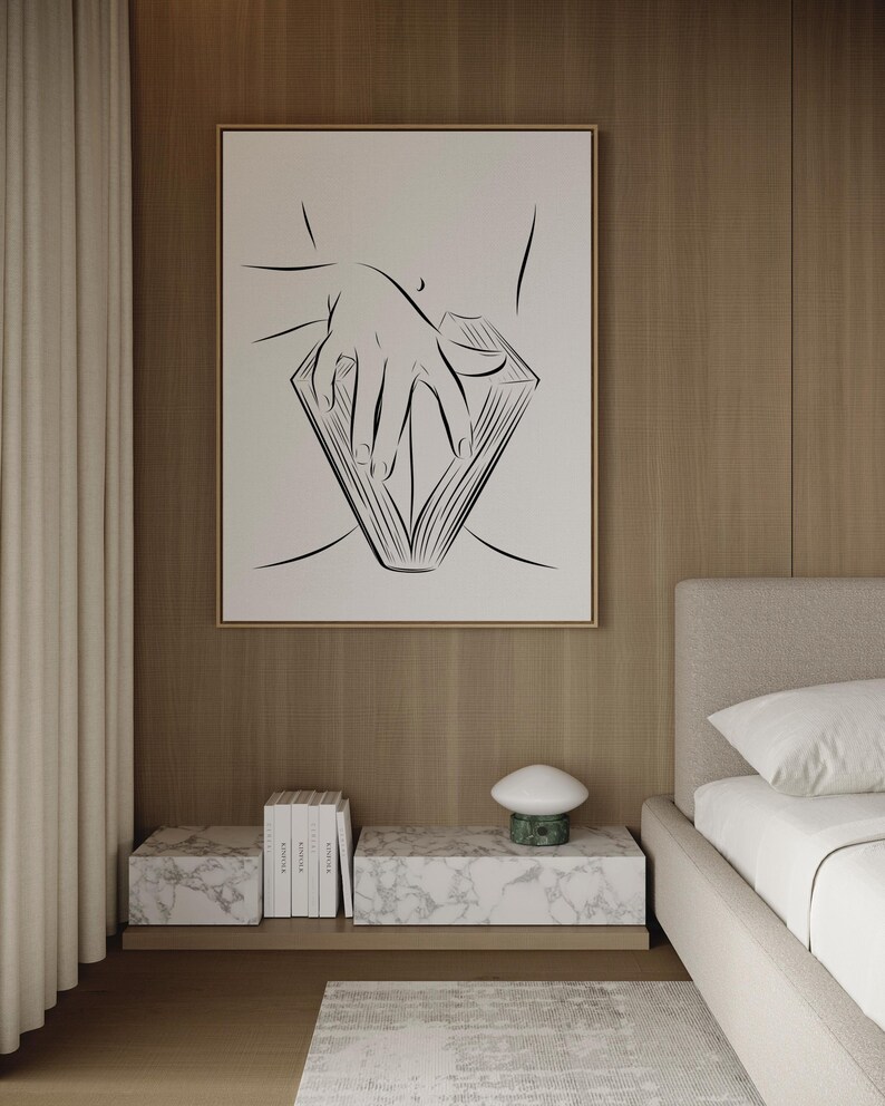 May include: A framed black and white line art print depicting a hand holding an open book. The artwork hangs above a marble-topped nightstand with books and a mushroom-shaped lamp. The room has a neutral color palette with wood paneling and a beige bed.