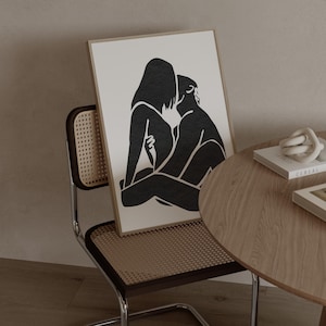 May include: A framed black and white art print depicting two figures in an embrace. The artwork is displayed on a chair with a woven back and seat. A wooden side table with books and a decorative knot is next to the chair.