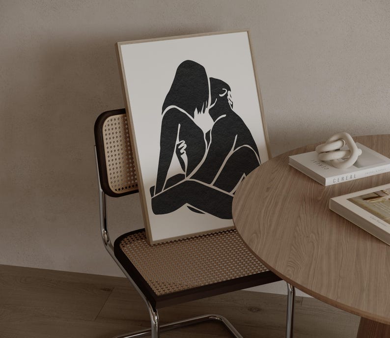 May include: A framed black and white art print depicting two figures in an embrace. The artwork is displayed on a chair with a woven back and seat. A wooden side table with books and a decorative knot is next to the chair.