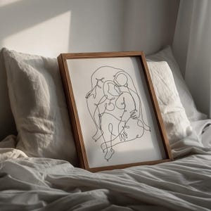 May include: A framed artwork featuring a minimalist line drawing of two figures embracing. The artwork is in a wooden frame and rests on a bed with white linens. The drawing is in black against a white background.