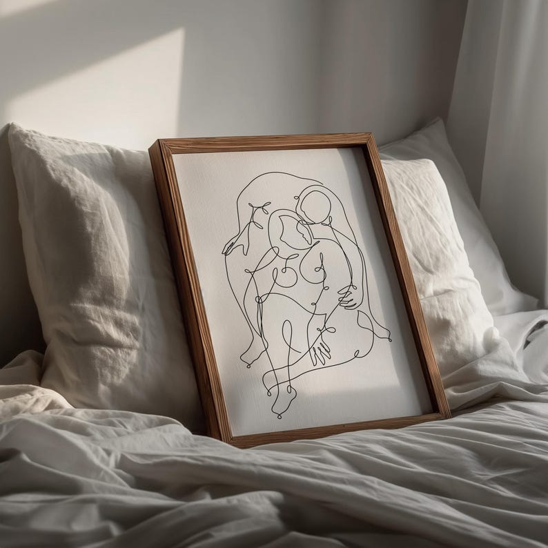 May include: A framed artwork featuring a minimalist line drawing of two figures embracing. The artwork is in a wooden frame and rests on a bed with white linens. The drawing is in black against a white background.