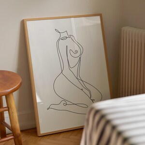 Puede incluir: Una impresi&oacute;n enmarcada con un dibujo lineal minimalista de una figura sentada. La obra de arte est&aacute; en negro sobre un fondo crema, dentro de un marco de madera marr&oacute;n claro. La obra de arte se apoya en una pared en una habitaci&oacute;n con un taburete de madera.