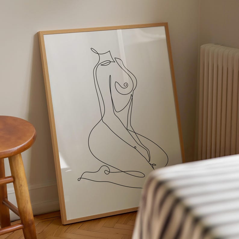 Puede incluir: Una impresi&oacute;n enmarcada con un dibujo lineal minimalista de una figura sentada. La obra de arte est&aacute; en negro sobre un fondo crema, dentro de un marco de madera marr&oacute;n claro. La obra de arte se apoya en una pared en una habitaci&oacute;n con un taburete de madera.
