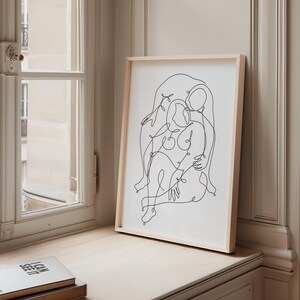 May include: A framed art print featuring a minimalist line drawing of two figures embracing. The artwork is rendered in black lines on a white background, set within a light wood frame. The print is displayed on a light-colored surface near a window.