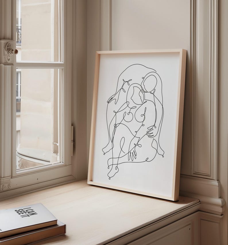 May include: A framed art print featuring a minimalist line drawing of two figures embracing. The artwork is rendered in black lines on a white background, set within a light wood frame. The print is displayed on a light-colored surface near a window.
