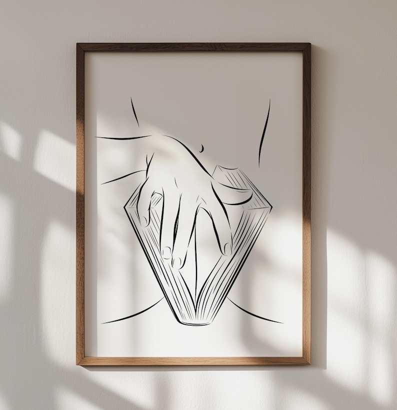 May include: A framed art print featuring a minimalist line drawing. The artwork depicts a hand resting on an open book, with the book's pages forming a diamond shape. The drawing is in black against a white background, encased in a brown wooden frame.