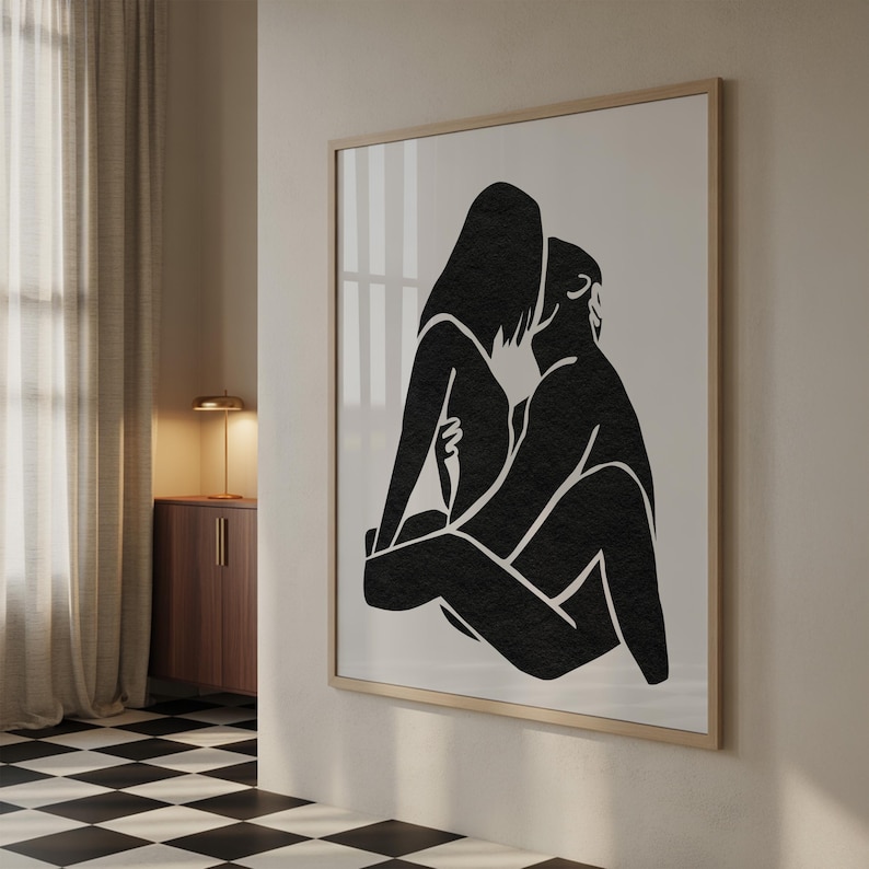 May include: Black and white art print featuring a minimalist illustration of two figures embracing. The artwork is framed in a light wood frame and hangs on a neutral-colored wall. The figures are depicted in a seated position.
