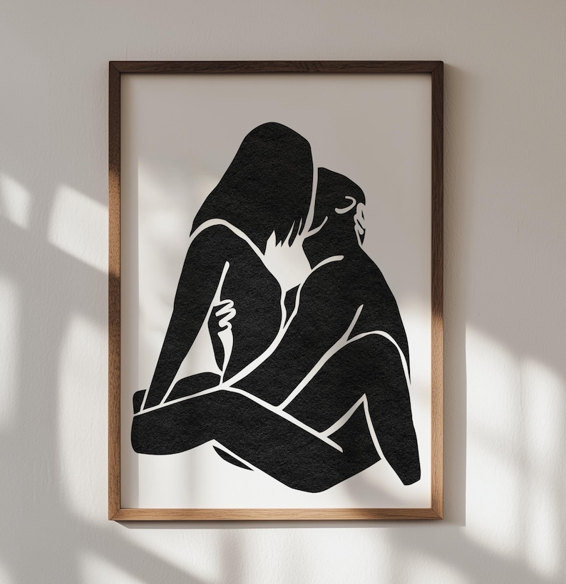 May include: Black and white art print featuring a minimalist illustration of two figures embracing. The artwork is framed in a brown wooden frame and mounted on a white background. The figures are depicted in a simple, abstract style.