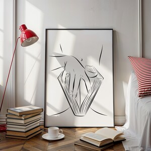 May include: Black framed art print featuring a minimalist line drawing of a hand holding an open book. The artwork is in black and white, with a simple yet elegant design. A red lamp and a stack of books are in the background.