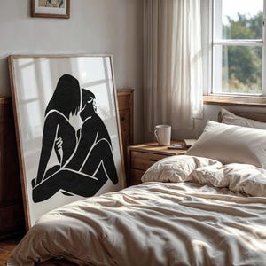 May include: A framed art print featuring a black and white abstract illustration of two figures embracing. The artwork is displayed in a bedroom setting, leaning against a wooden wall. The room has a neutral color palette.