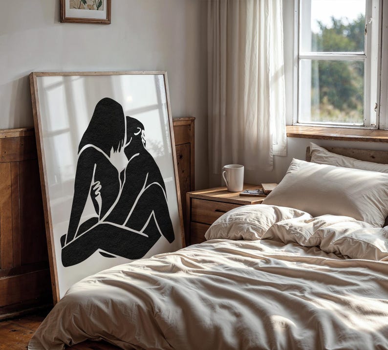 May include: A framed art print featuring a black and white abstract illustration of two figures embracing. The artwork is displayed in a bedroom setting, leaning against a wooden wall. The room has a neutral color palette.