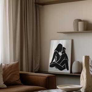 May include: A framed black and white art print depicting two figures in an embrace. The artwork is displayed on a shelf with decorative vases, set against a neutral-toned wall and a brown leather sofa. The room has a minimalist aesthetic.