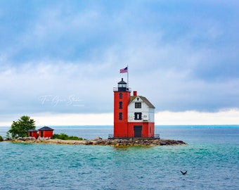 May include: A red and white lighthouse with a small house attached stands on a small island in a body of water. The lighthouse has a flagpole with an American flag flying from it. The water is a light blue color and the sky is a light blue color with some clouds.